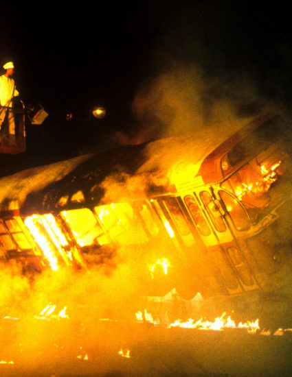 Un autobus sur une broche géante rotative, prend feu, pendant qu'un comédien vêtu d'un costume de cuisinier surveille la "cuisson"