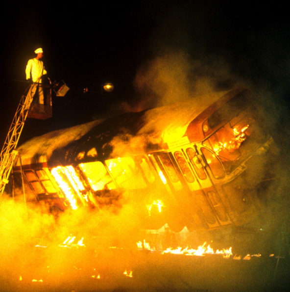 Un autobus sur une broche géante rotative, prend feu, pendant qu'un comédien vêtu d'un costume de cuisinier surveille la "cuisson"