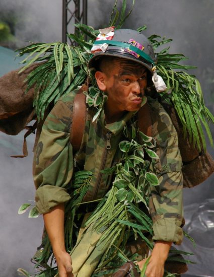 Un homme, vêtu d'une treillis militaire, casque sur la tête sur lequel est accroché un paquet de cigarette