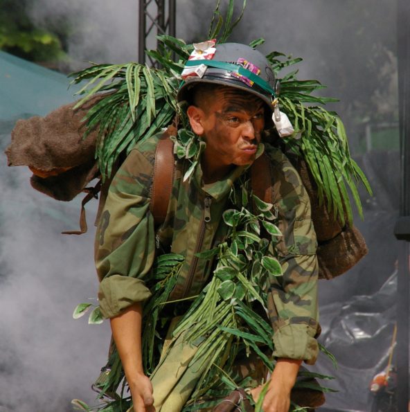 Un homme, vêtu d'une treillis militaire, casque sur la tête sur lequel est accroché un paquet de cigarette