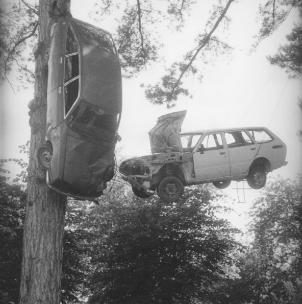 Une voiture est suspendue au tronc d'un grand arbre. Une autre voiture derrière est suspendue dans les branches et flotte au dessus de la terre