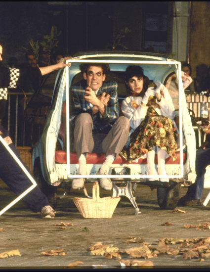 Le couple de comédien du Roman Photo est dans une voiture fiat, coupée en deux pour laisser voir l'intérieur. Les techniciens sont sur le côté de l'image et tiennent le cadre qui représente la scène du Roman photo